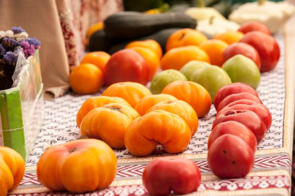 Red, green, and orange tomatoes are shown on a table with a red and white tablecloth at a local Farmers Market. There are other vegetables in the distance. 