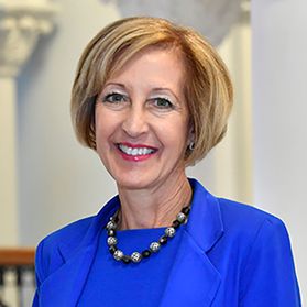 Donna Peduto smiles with short hair, red lipstick, a blue blouse and jacket and bulb necklace