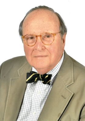 Headshot of retired WVU administrator Robert DiClerico. He is pictured against a light background wearing a tan sport coat over a white patterned dress shirt and a dark striped bowtie. He wears round framed glasses and has gray hair.  