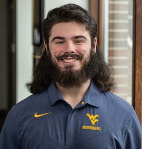 Headshot of WVU Mountaineer mascot candidate Justin Waybright. He is standing in front of a window and wearing a navy blue WVU-branded shirt. He has a long black mullet, a long black bear and a black handlebar mustache. 