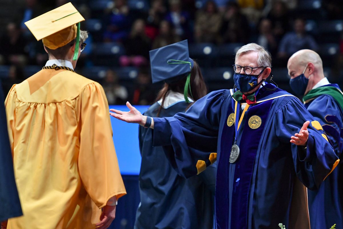 man in graduation regalia greets with open arms a person in cap and gown