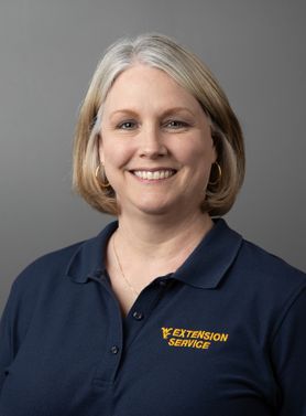 A person with chin length hair smiles in front of a gray backdrop while wearing a navy blue polo shirt with WV Extension Service on the left side.