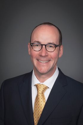 Headshot of WVU Medicine chief Albert Wright. He is pictured against a gray background and is wearing a dark colored suit with a white dress shirt and gold, dotted tie. He has very short dark hair and round-framed glasses. 