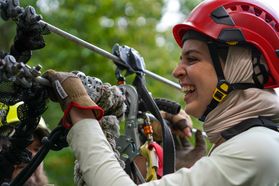 A woman prepares to ride the length of a zip line while wearing a red helmet and smiling. 
