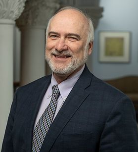 A portrait shot of Paul Kreider, pictured smiling. He has a white beard and is wearing a suit and tie.