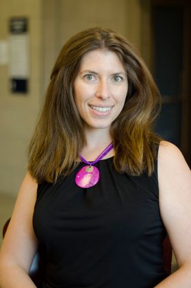 Photo of a woman with long light brown hair smiling, wearing a large magenta necklace