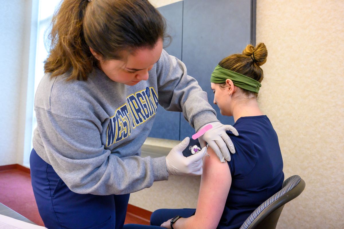 A nurse dressed in a West Virginia University sweatshirt and navy blue scrubs administers a shot to a female student. 