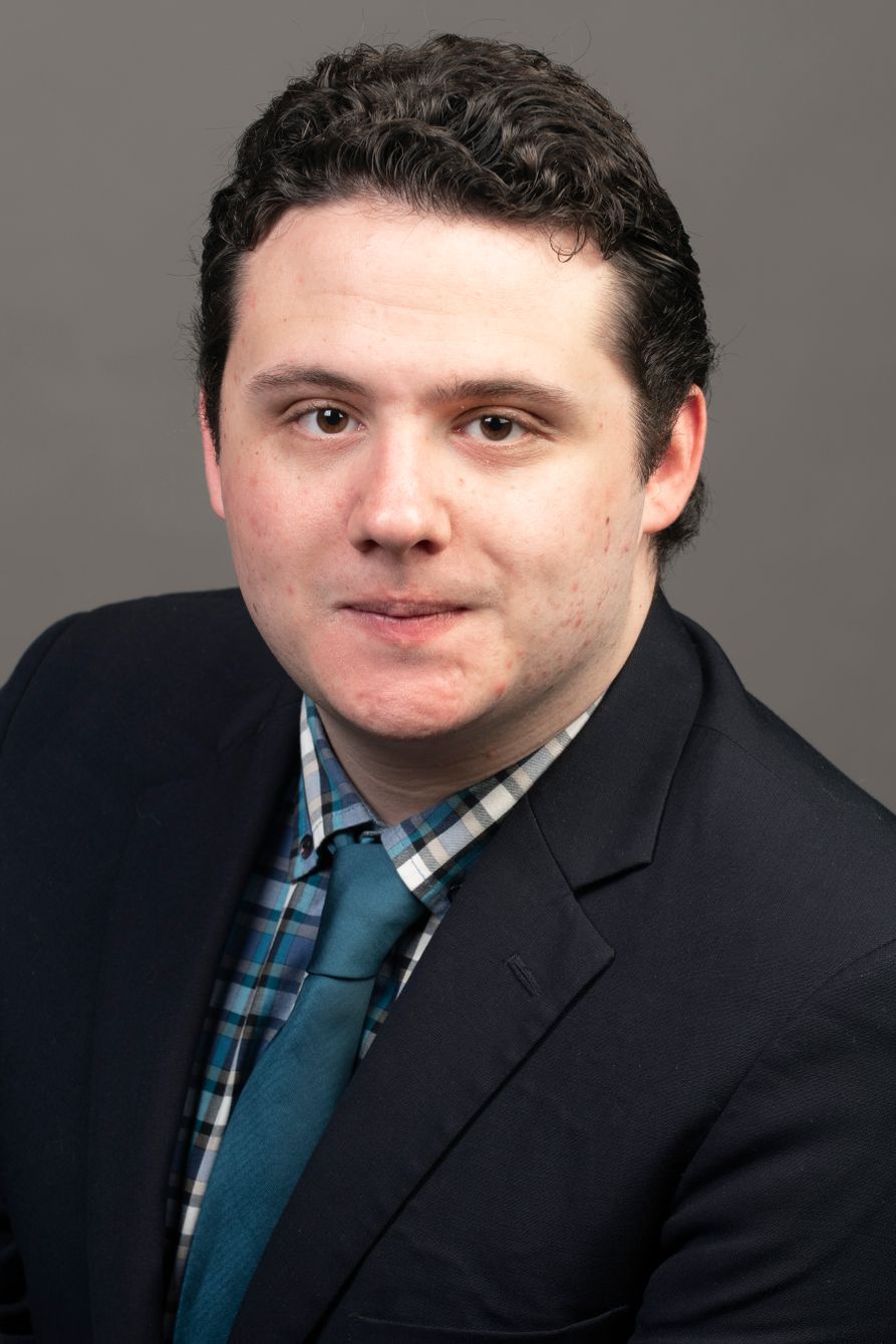 man with short dark hair wearing a plaid shirt, tie and dark suit jacket