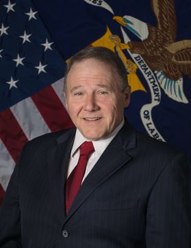 Man standing in front of American flag with dark blue suit, white shirt and red tie