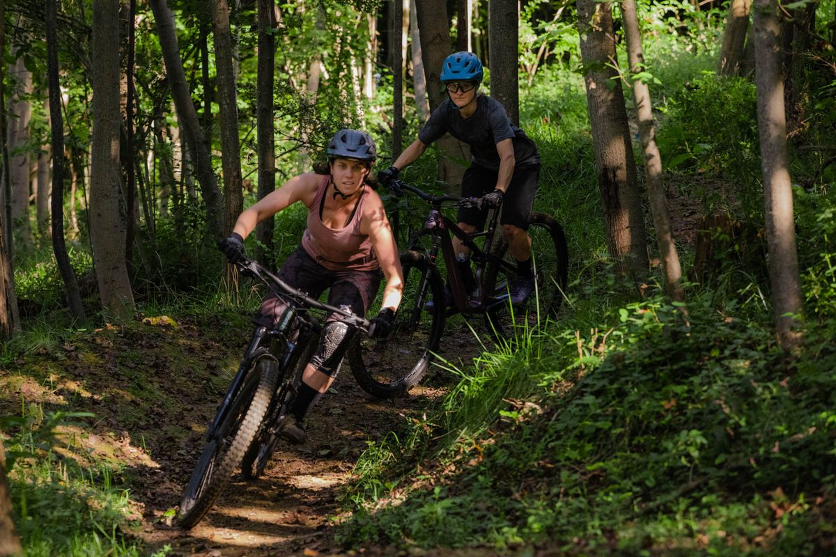 Two picture are on bikes while riding down a dirt trail between trees. Both are wearing blue helmets. The person in the front has on a pink short sleeve shirt. The person in the back is in a dark shirt.