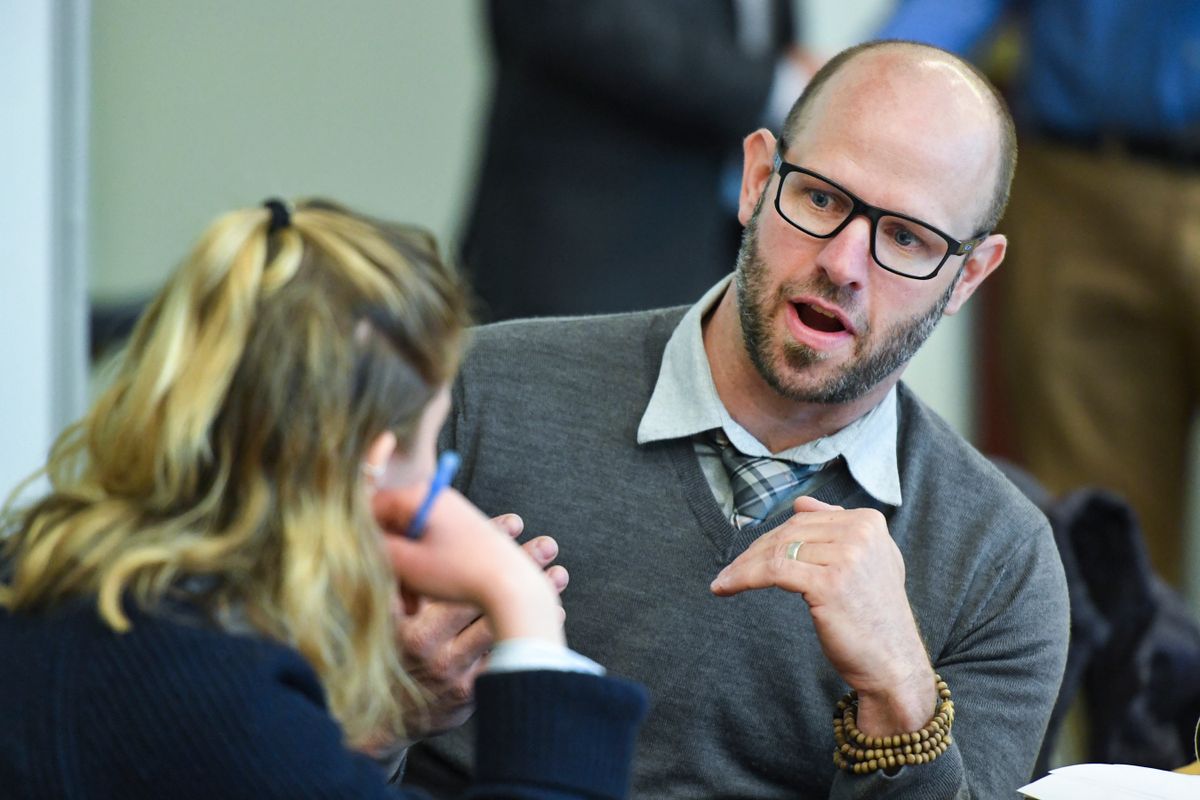 Man in a grey sweater and glasses explaining something to a blonde woman with her back to us holding a pen.