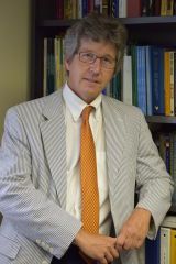 A gentleman in a grey suit and orange tie and grey hair wearing glasses leans against a stocked bookshelf for a photo.