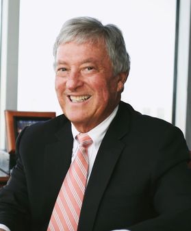 A man wearing a black suite jacket over a white dress shirt with a pink and tan stripped tie. He has gray hair.