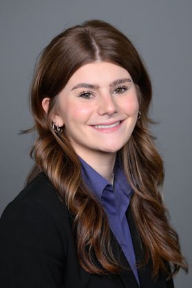 Headshot of WVU Bucklew Scholar Samantha Guenther. She is pictured against a gray background wearing a black jacket over a blue blouse. She has long, wavy Auburn hair.  