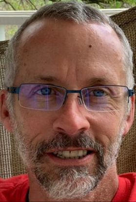 A headshot of WVU researcher Tim Driscoll. He is pictured inside wearing a red shirt and glasses. He has receding gray hair and a gray beard. 