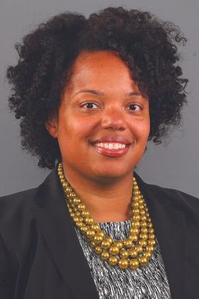 Headshot of Natasha Oakes. She is pictured against a gray background. She is wearing a black jacket over a black and white patterned blouse. She is wearing a gold beaded necklace. He has short, curly black hair 