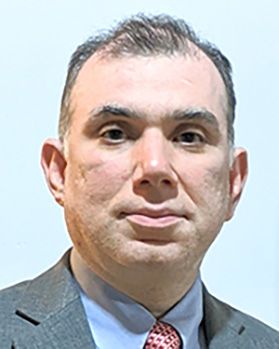 Headshot of WVU expert Khalid Kamal. He is pictured against a light blue wall and is wearing a gray suit with a light blue dress shirt and red patterned tie. He has thinning dark colored hair. 