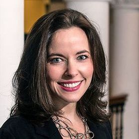 Headshot of Dana Voelker. She has long, brown hair and is wearing a black top with a statement necklace. She is pictured inside with columns in the background. 