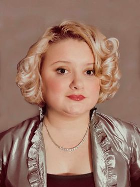 Headshot of WVU musical theatre student Cadee Mae Green. She has short, blonde, curly hair and is wearing a silver jacket over a black blouse. 