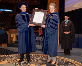 Scott Widmeyer and Provost Maryanne Reed stand on the WVU Commencement stage. Reed is presenting Widmeyer an honorary degree in a large wooden frame.