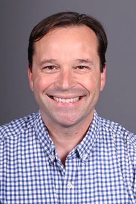 Headshot of WVU researcher Daniel Renfrew. He is pictured against a gray background wearing a blue checked dress shirt. He has short, dark hair and a big smile. 