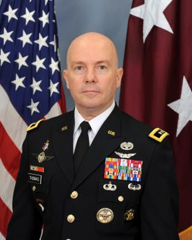 A bald man in uniform with several service medals in front of the American Flag