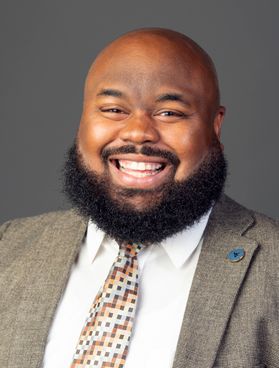 This is a portrait of Javier McCoy. He is smiling in front of a gray backdrop and is wearing a gray suit jacket, spotted gold and brown tie and white button-up shirt. He has a dark beard.