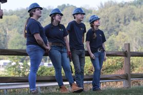 group of young men and women lean on a wooden fence in matching hard hats and t-shirts