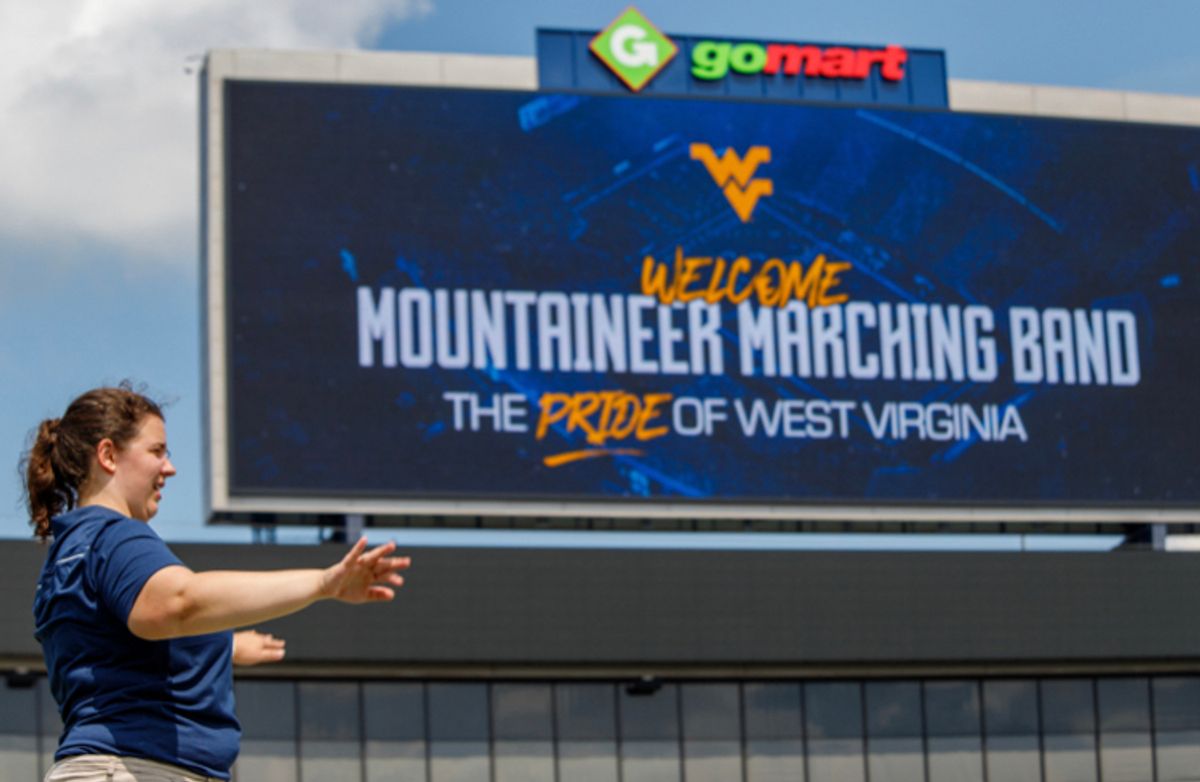A girl's profile standing all the way to the left with a huge WVU Mountaineer Marching Band billboard in the background