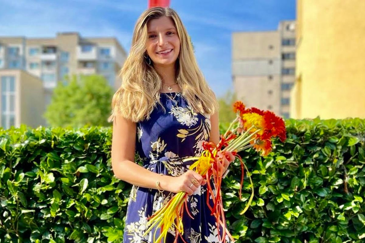 Photo of WVU alumna Giana Loretta standing in front of a green hedge holding a large bouquet of red and yellow flowers. She has long blonde hair and is wearing a navy blue floral dress. 