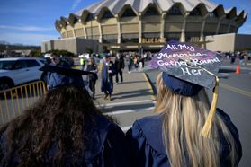 Two graduates wearing navy blue caps and gowns are show from the back walking to Coliseum. The one on the left has long dark hair. The one on the right has long blonde hair is wearing a mortarboard with the words 'All My Memories Gather Round Her' on it.
