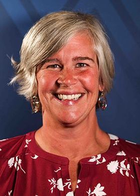 Headshot of WVU Extension specialist Gina Wood. She is pictured in front of a blue background and is wearing a red and white floral shirt. She has short blonde hair and wears dangling gold earrings. 