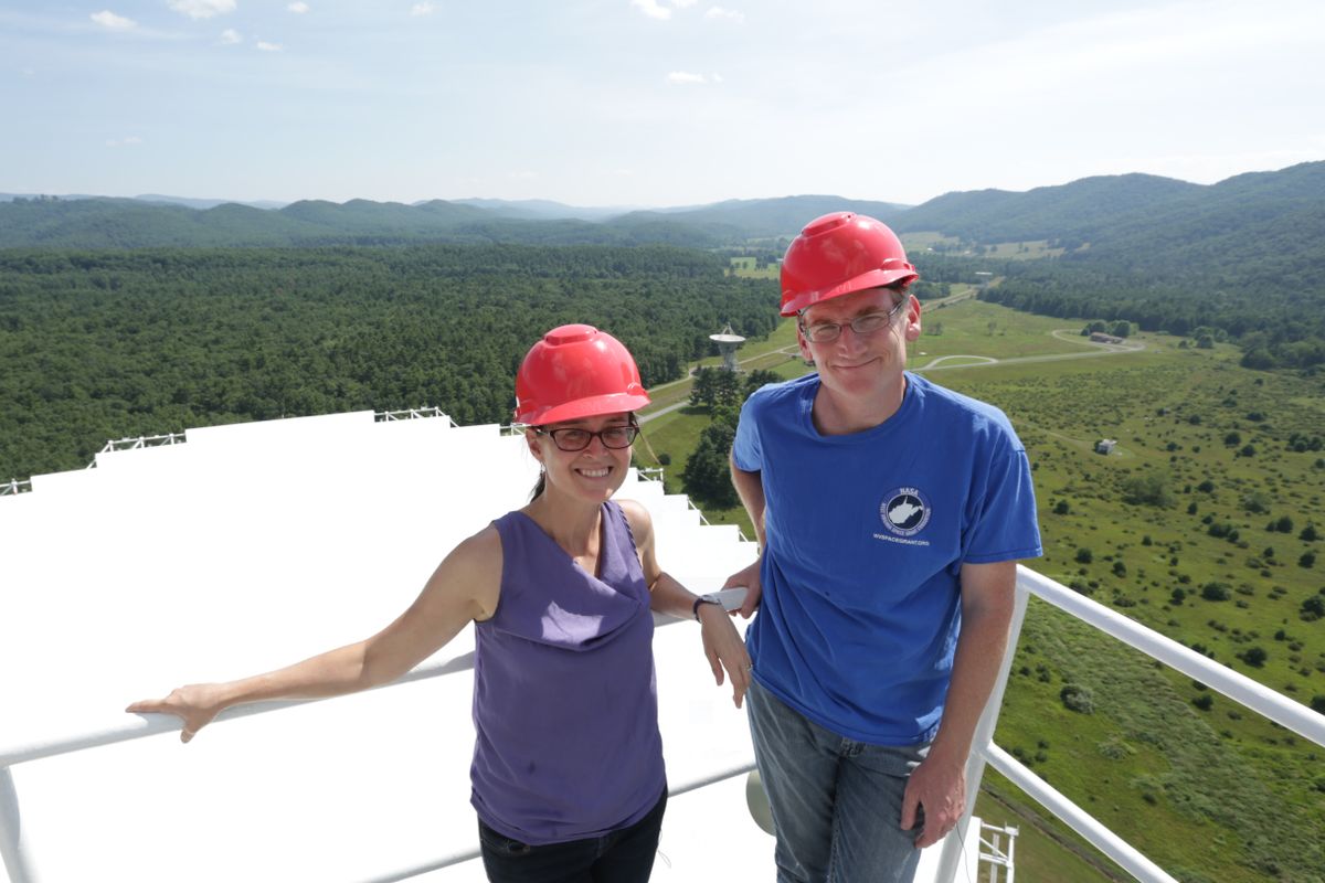 (Left) A woman in a purple tank top and a red hard hat and (Right) a man in a blue t shirt and a red hard hat pose on a gate in front of mountains.