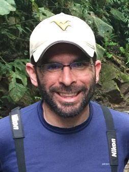 Headshot of WVU researcher Chris Rota. He is standing outside wearing a blue t-shirt and a white baseball cap. He is wearing glasses and has a dark colored beard. 