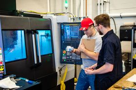 Two male students work stand near industrial equipment inside the WVU Lane Innovation Hub. One student is holding a clip board and wearing a red hat while the other student is dressed in a dark colored shirt and wears glasses
