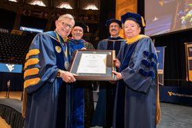three men stand with a woman (right) holding a diploma on stage in cap and gown