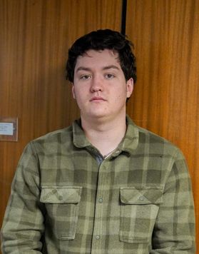 Headshot of WVU student Justin Grimes. He is standing in front of a wood paneled wall wearing a green plaid flannel shirt. He has short black hair. 
