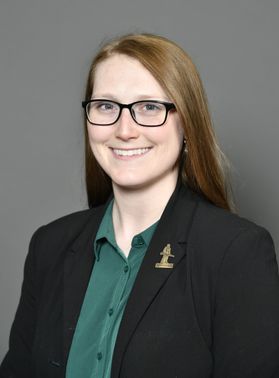 A woman with straight brown hair and glasses smiles while wearing a green shirt and black blazer
