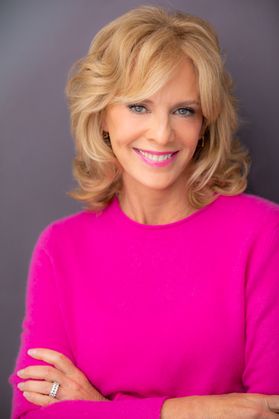 Headshot of Sandy Bainum, a WVU alumna who found success in the performing arts. She is pictured against a gray background with her arms crossed in front of her. She is wearing a bright pink top and has shoulder-length blonde hair. 