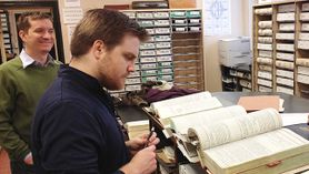 Two male WVU law students wearing a green shirt and blue shirt look through records at a courthouse surrounded by books.