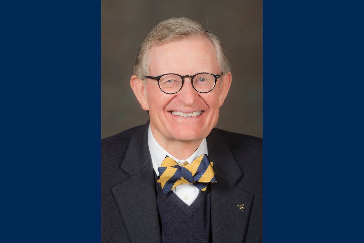 This is a portrait of WVU President Gordon Gee on a blue background. Gee is wearing a dark jacket, vest, gold and blue tie, and black-rimmed glasses.