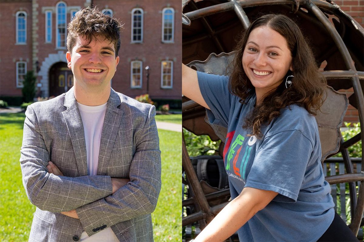 A young man with curly dark hair smiles outside with his arms crossed, wearing a suit jacket, while a girl with long  dark curly hair wears a blue shirt and smiles