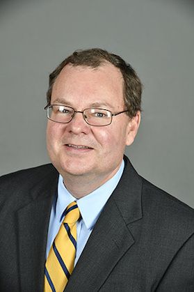 A man in front of gray wall looks at the camera while wearing wire rimmed glasses. His suit is dark gray. His shirt is blue. His tie is mostly gold with a blue stripe.