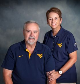 Photograph of Dr. Jo Ann and David Shaw. They are pictured in front of a gray background and both are wearing navy blue WVU-branded gold shirts. Jo Ann has short red hair and David has gray hair and a full gray beard. 