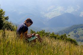 A photograph of a male WVU student sitting on a hillside writing in a book. The view of mountains in the distance is expansive. 