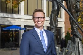 Headshot of WVU student Matthew Hudson standing in front of the Mountainlair wearing a blue suit and tie. 
