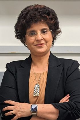 Headshot of WVU professor Shikha Sharma. She is standing against a beige background and is wearing a black jacket over a rust-colored blouse. She is also wearing a large pendent and an Apple watch. She has short curly dark hair and has her arms crossed. 