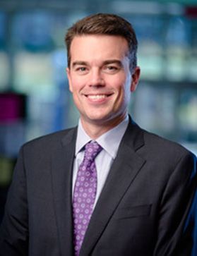 Headshot of WVU dean Josh Hall. He is pictured inside with a blurred background behind him. He is wearing a black sport coat over a white dress shirt and with a purple tie. He has short brown hair. 