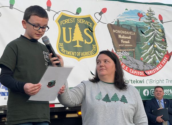 Ethan Reese is shown in a green shirt and long dark sleeves on the left of the frame holding his winning essay. His mom, in a gray sweatshirt with green trees, holds a microphone in front of a banner for the U.S. Capitol Christmas Tree.