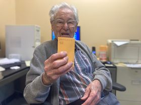 WVU researcher Mannon Gallegly poses in an office holding a small yellow envelope of tomato seeds for anew variety that he's just released. He is wearing a gray cardigan over a plaid shirt. He has silver hair and wears glasses. 